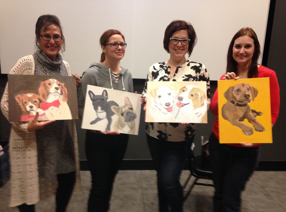 Four women posing with paintings they created of their pets.